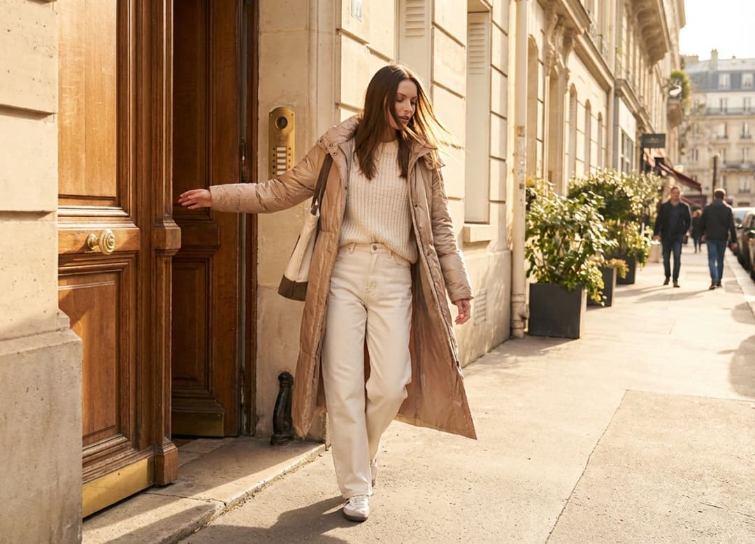 Mannequin en doudoune longue beige Misciano sortant d’un immeuble, capturée en mouvement.