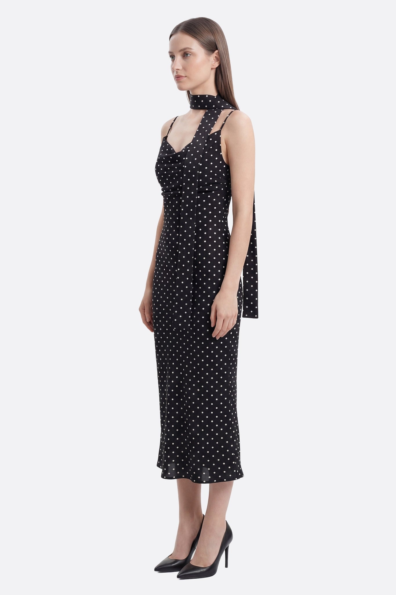 Model in profile wearing a black midi dress with white polka dots and removable strap at the neck