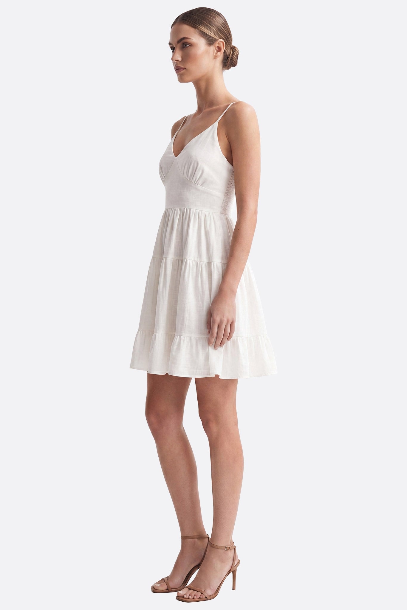 Model side view wearing a white short dress with thin straps, smocked back and ruffled skirt