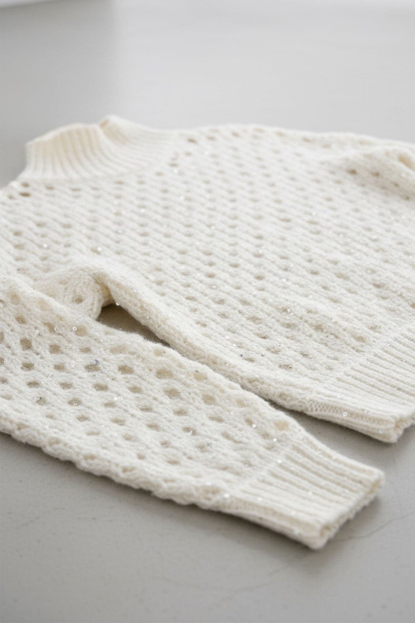 Nahaufnahme des weißen Lochmuster-Kaschmirpullovers mit Stehkragen, die die Pailletten für einen Hauch von Eleganz hervorhebt...