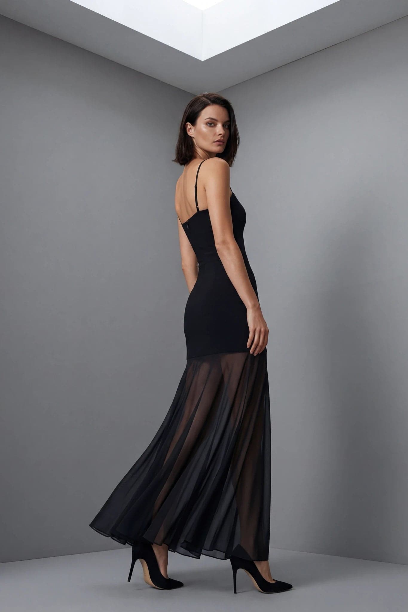 Model trägt von hinten ein langes transparentes schwarzes Kleid mit feinen Trägern und fließendem Rock
