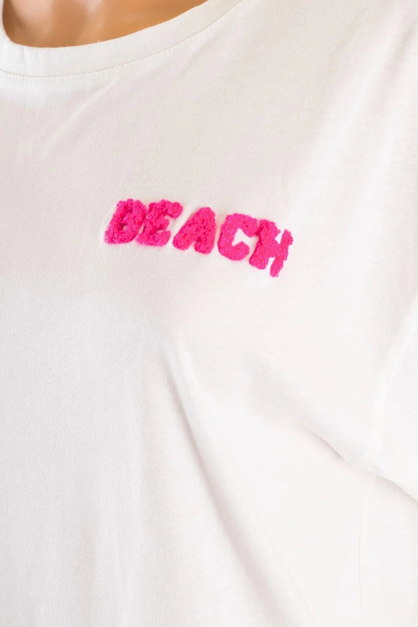 Деталь белой футболки oversize из мягкого хлопка, раскрывающая вышивку BEACH, идеальную для повседневного и утонченного стиля.