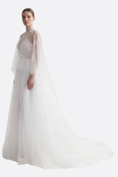 
Détail du corsage brodé de la robe de mariée en tulle, avec jupe fendue et cape amovible en arrière-plan.



